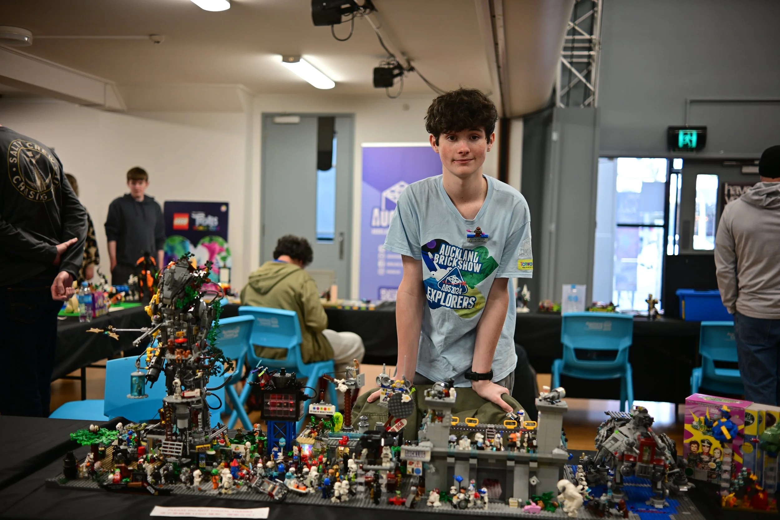 A boy with curly hair wearing a gray T-shirt that says 'Auckland Brick Show Explorers' stands behind a table displaying detailed LEGO structures and models at a LEGO exhibition or competition.