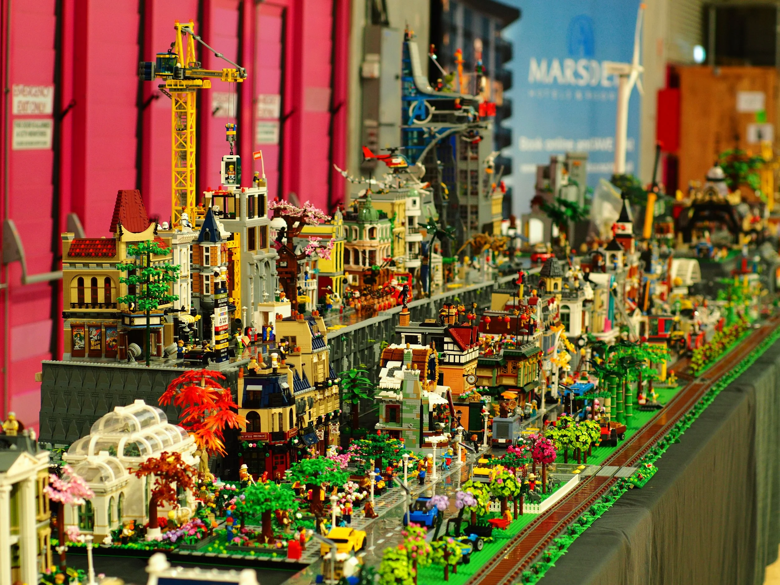 A detailed LEGO cityscape on a display shelf featuring numerous buildings, figures, trees, vehicles, and a construction crane, set against a background of colorful panels and a blue sign.