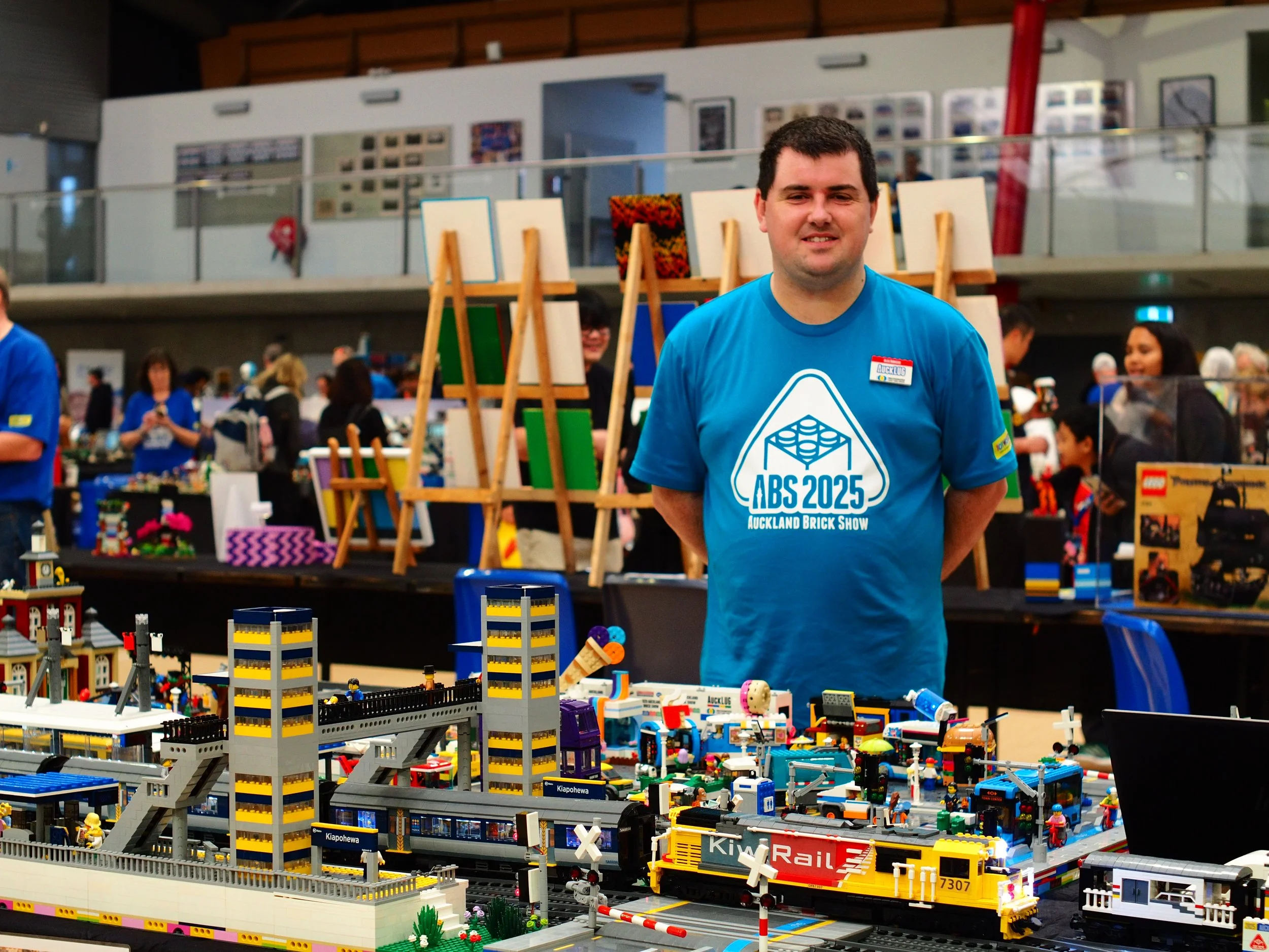 A man wearing a blue T-shirt and a name tag at an exhibition table covered in LEGO models, including a large cruise ship and a train, with many other people and displays visible in the background.
