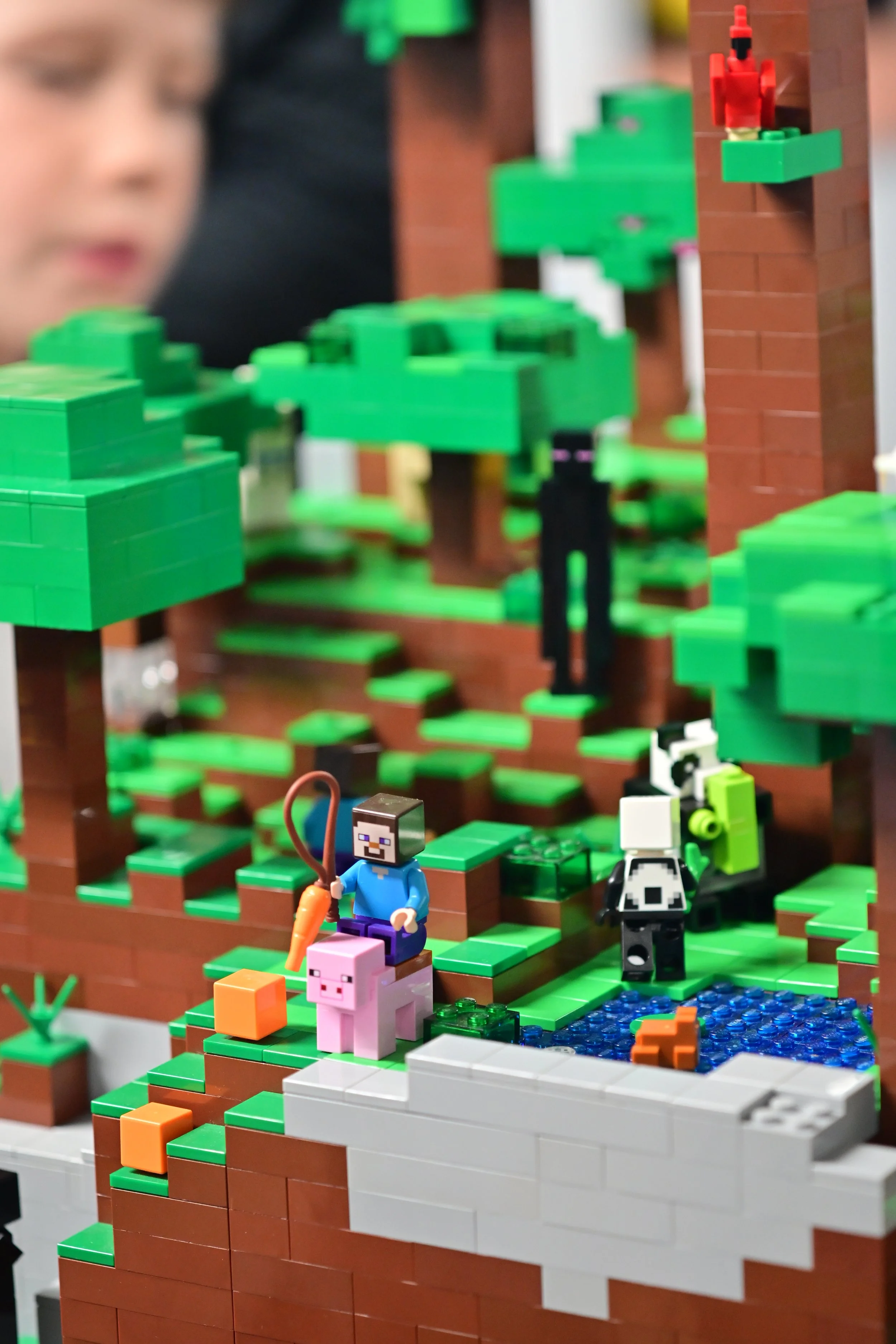 A detailed Lego scene featuring pixelated characters from Minecraft, including a character holding a carrot and a pig, set against a backdrop of Lego trees, a pond, and a brick structure.