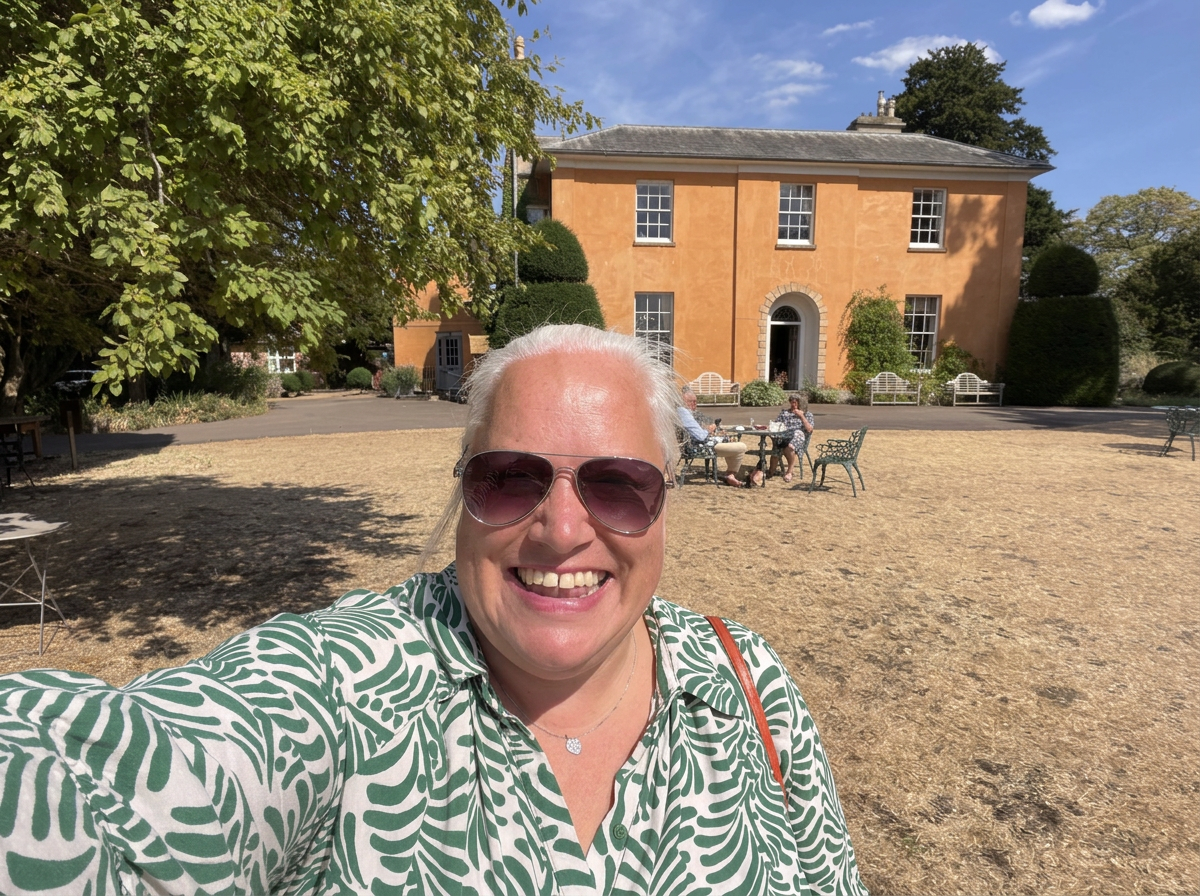 white woman in sun glasses smiling at the camera.  In the background is a large house and a lawn with people sitting at a table with drinks