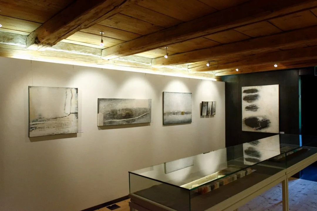 Kunstwerke in einem Museumsraum, schwarz-weiße abstrakte Gemälde an einer Wand, Holzdecke und Glaskästen mit Objekten im Vordergrund.