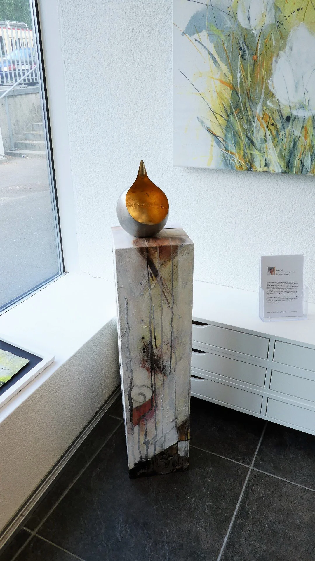 Kunstwerk auf einem hohen Podest, eine metallene, ovale Skulptur mit einer spitzen Spitze an der Oberseite, im Kunstmuseum. Im Hintergrund eine abstrakte Wandmalerei mit Linien und farbigen Akzenten.