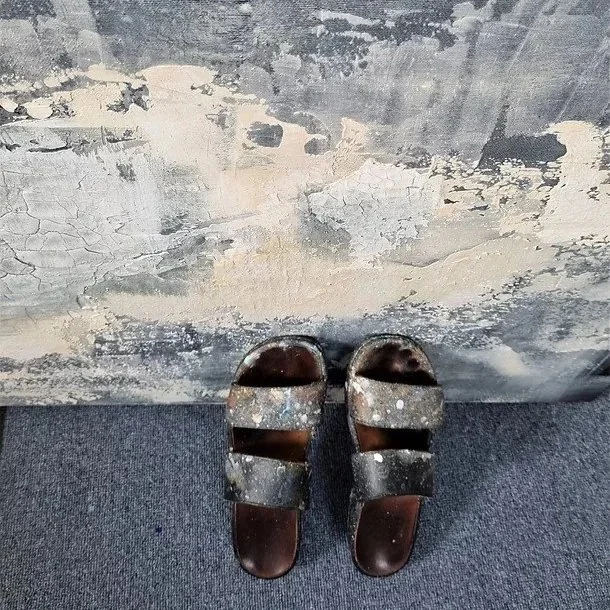 Zwei schwarze, abgenutzte Sandalen stehen auf einer grauen, verschmutzten Oberfläche vor einer Wand mit abblätternder Farbe.