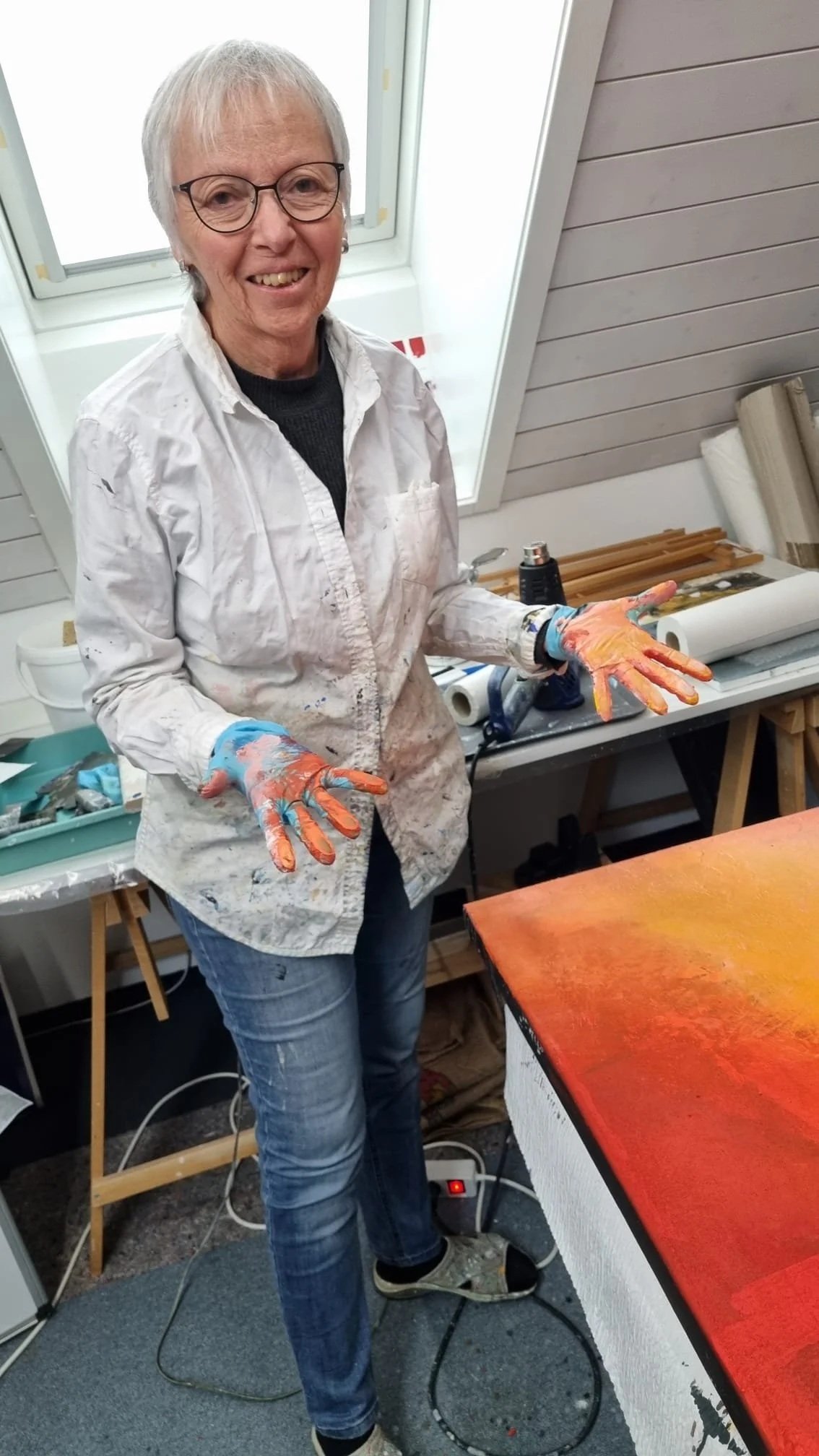 Eine ältere Frau in weißer Kunstjacke und blauen Jeans zeigt ihre bemalten Hände in einem Kunst-Workshop-Raum, umgeben von Malutensilien und einem Farbkunstwerk auf einem Tisch.