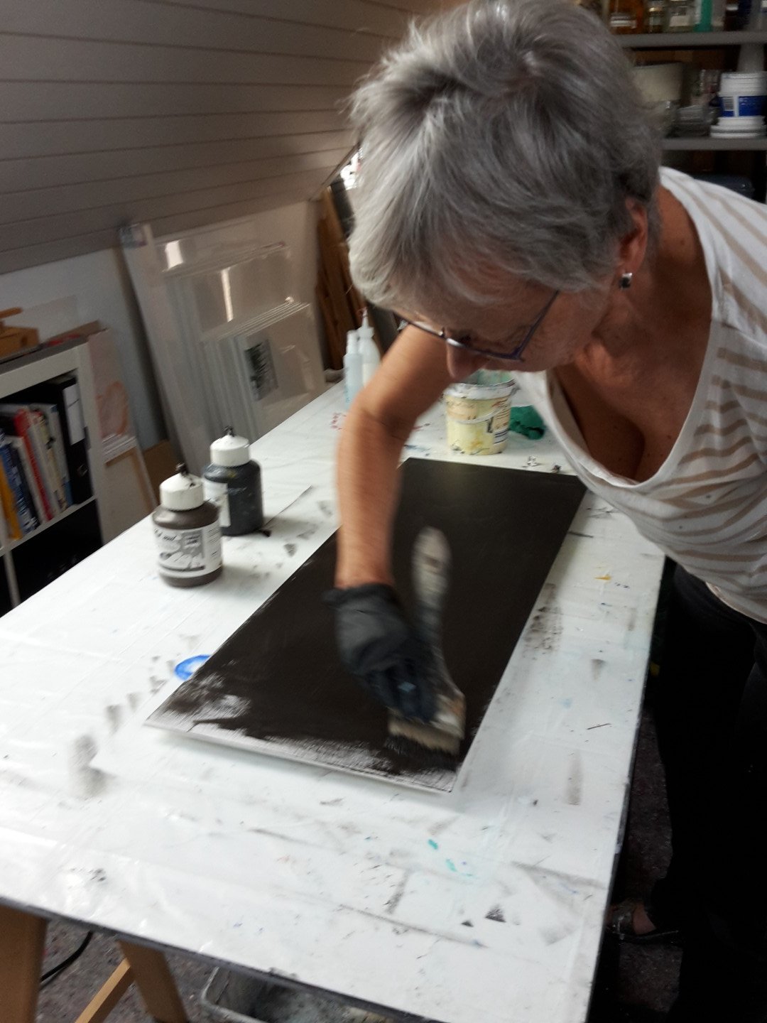 Eine ältere Frau trägt Handschuhe und streicht mit einem Pinsel über eine dunkle Fläche auf einem Tisch in einem Raum, der als Werkstatt oder Atelier aussieht.