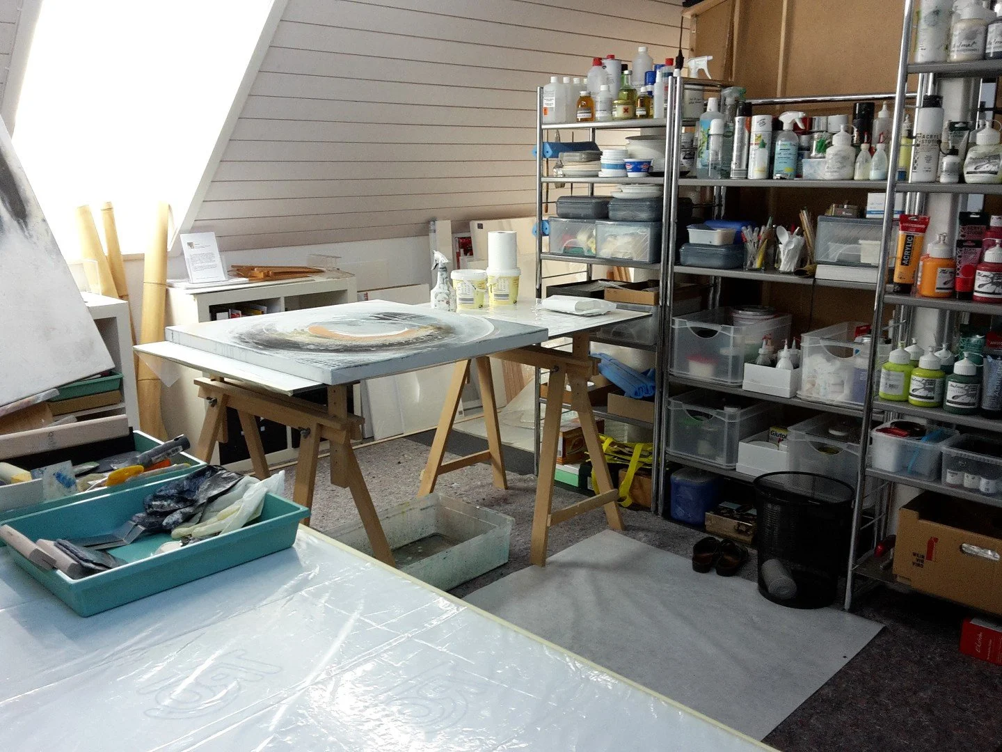 Künstleratelier mit Werkbänken, Regalen voller Farben und Materialien, Leinwänden und Arbeitsutensilien.