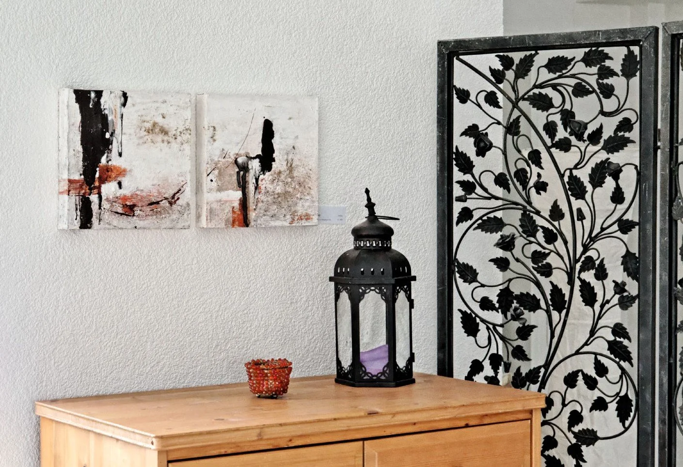 Eine Holztisch mit Laterne in schwarz und einer kleinen Kerze in einem roten Glas-Untersetzer. Über dem Tisch an der Wand hängen zwei abstrakte Gemälde. Rechts ist ein dekoratives Gitter mit einem Blattmuster.