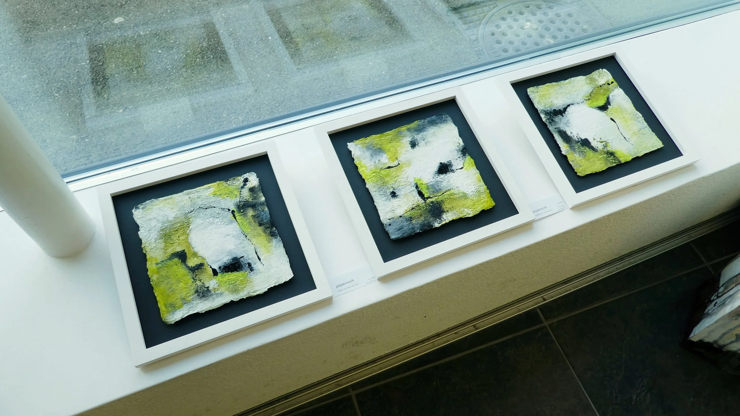 Drei abstrakte Gemälde einer Serie in Grün-, Weiss- und Schwarztönen, welche in einem Ausstellungsraum auf einer Fensterbank liegen.