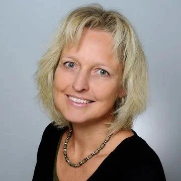 Eine Frau mit blonden, lockigen Haaren, die ein schwarzes Oberteil trägt und eine Halskette trägt, lächelnd vor hellem Hintergrund.