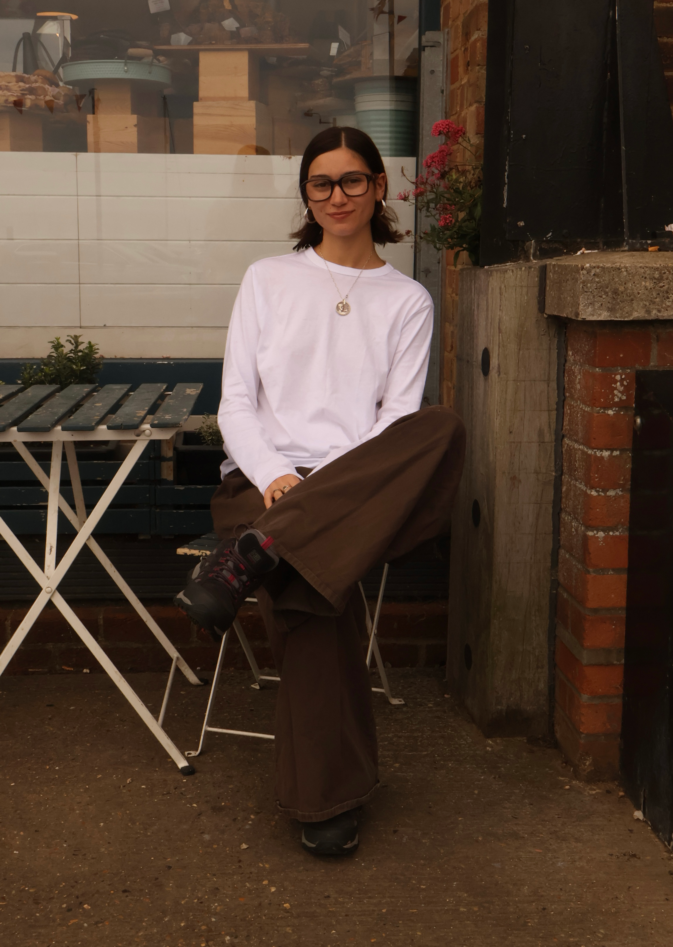 A young woman with shoulder-length dark hair, wearing glasses, a white long-sleeve shirt, and brown wide-leg pants, is sitting on a metal chair outside near a brick wall and window. She is smiling and looking at the camera, with one leg crossed over the other. There are plants, a folding table, and a window with visible indoor items in the background.