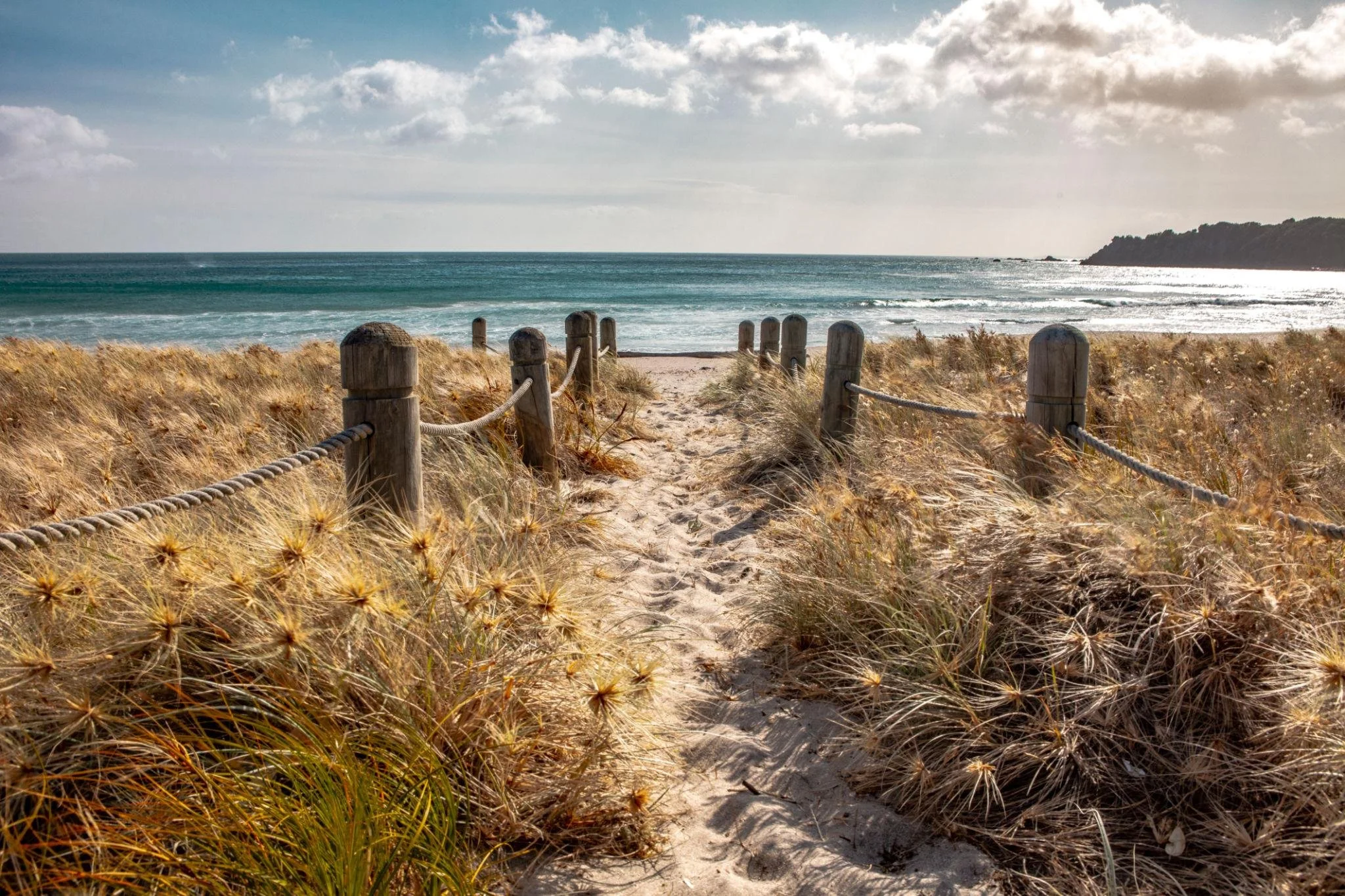 A sandy beach with tall grass leading to the ocean, with a wooden rope fence near the path, cloudy sky, and distant land on the horizon.
