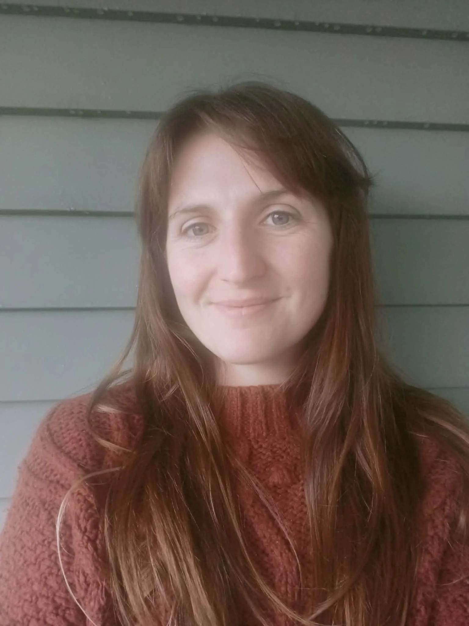 Meg O’Sullivan, Clinical Psychologist at Bay Psychological Services. A woman with long red hair wearing a red sweater, standing in front of a gray horizontal siding wall.