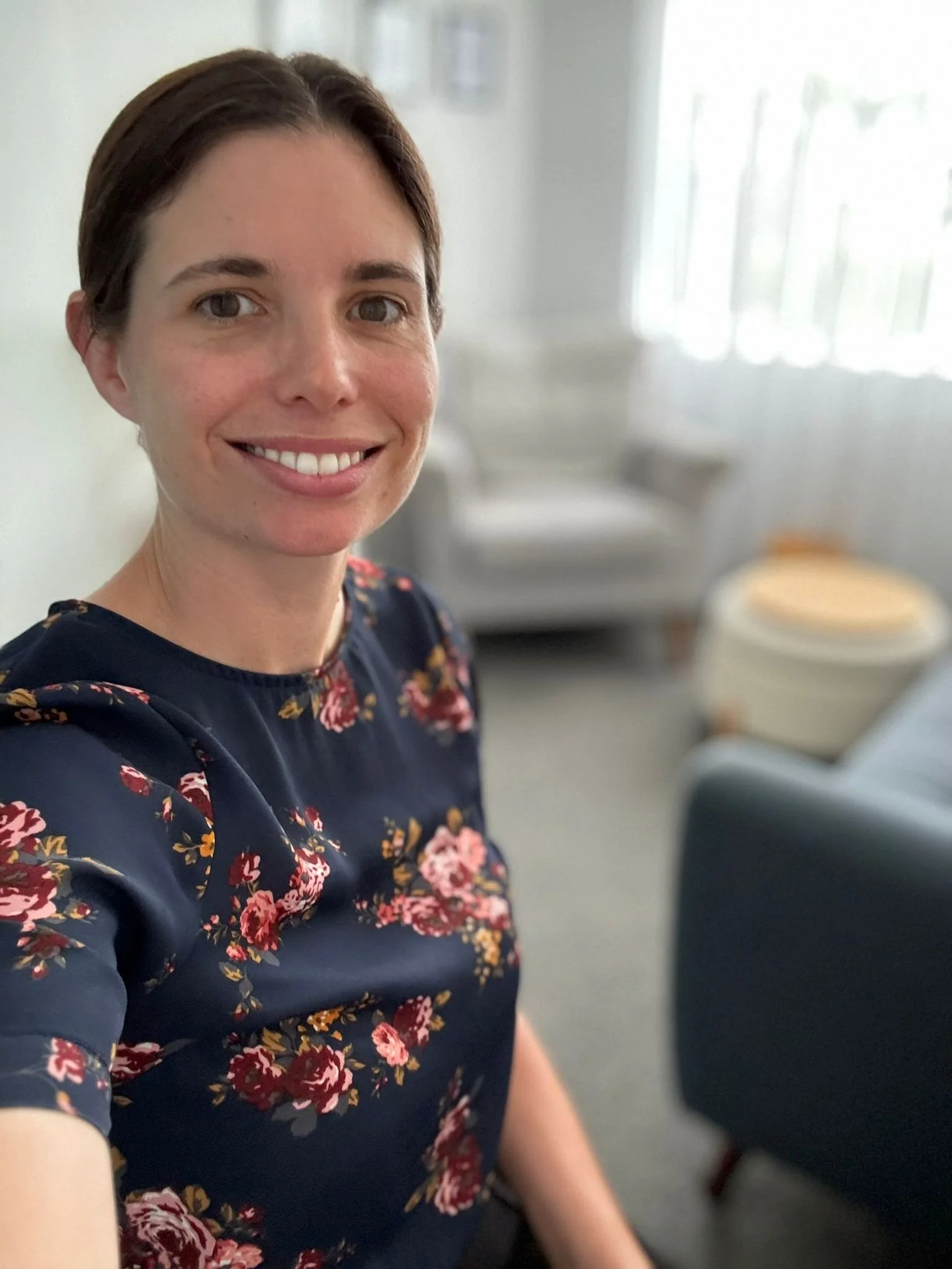 Amelia Ryan, Clinical Psychologist at Bay Psychological Services. A woman with short brown hair smiling, wearing a navy floral dress with pink and red flowers, sitting in a room with a window in the background.
