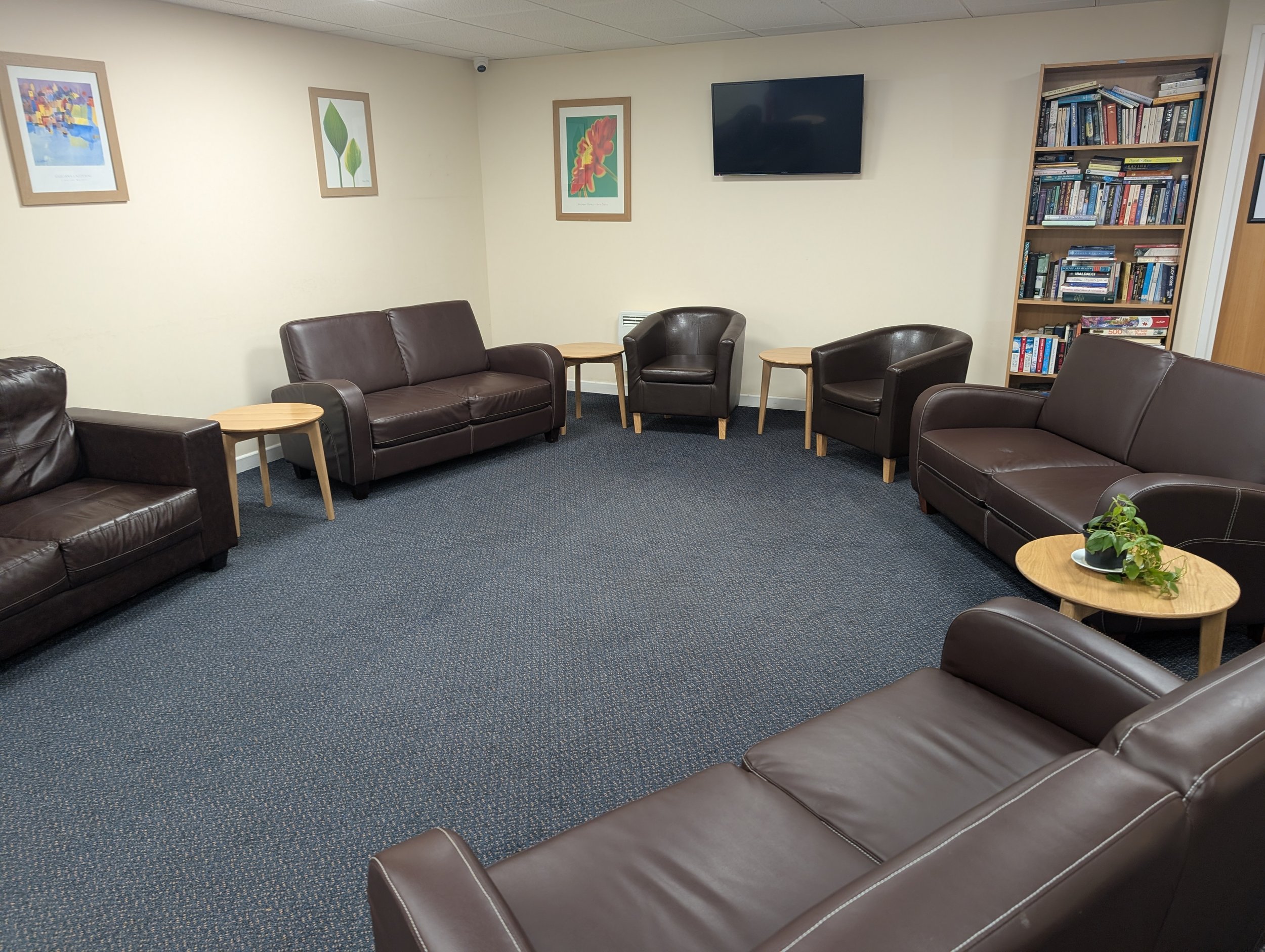 A waiting room with six dark brown leather sofas and three small wooden tables. The room has beige walls, a blue carpet, framed colorful artwork, a flat-screen TV, a wooden bookshelf filled with books, a plant on a side table, and a closed door.