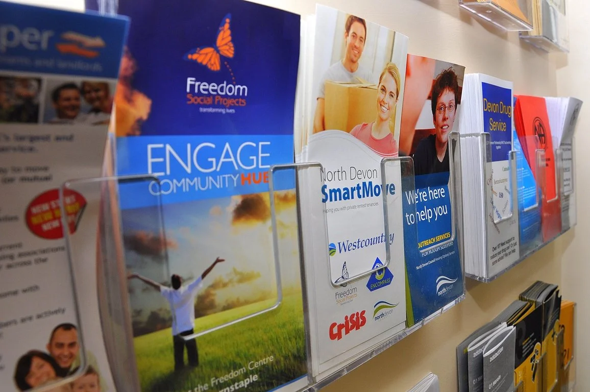 Brochures and flyers in a pamphlet rack promoting community engagement, social projects, and local services in North Devon.