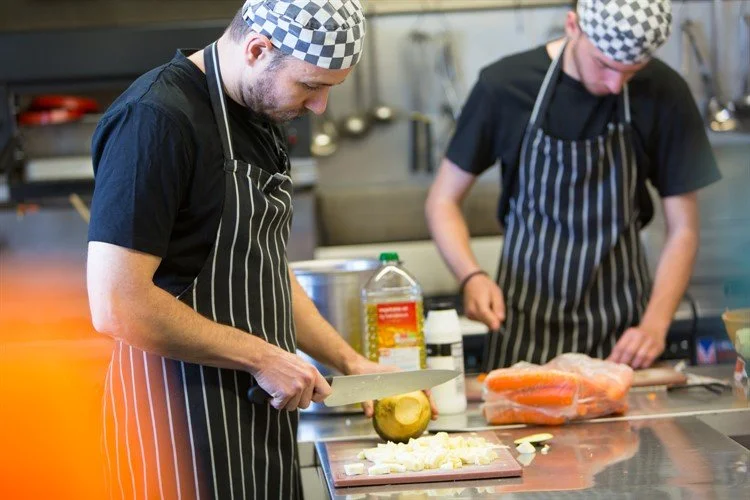Two chefs in black shirts and striped aprons preparing food in a professional kitchen. One is chopping a pineapple while the other is working with a salmon fillet.