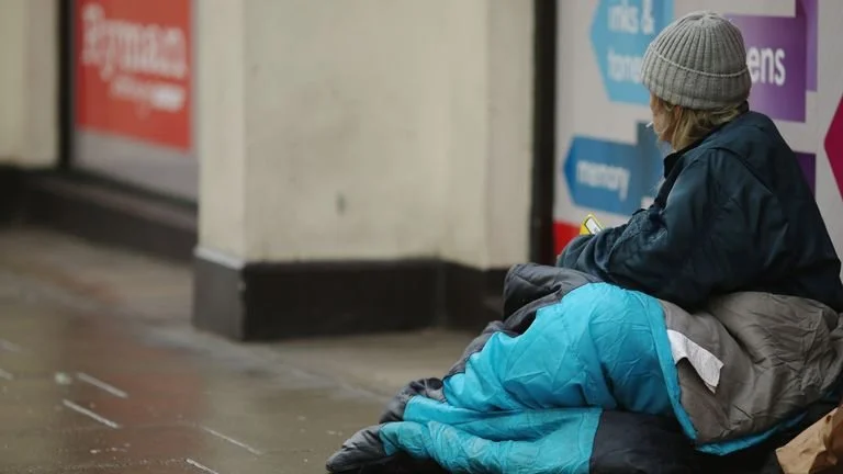 A person dressed in a blue jacket and gray beanie sitting on the ground near a wall with posters, looking at their phone.