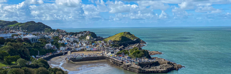 A coastal city with colorful buildings, a sandy beach, and a pier extending into the ocean, with hills in the background and partly cloudy skies.