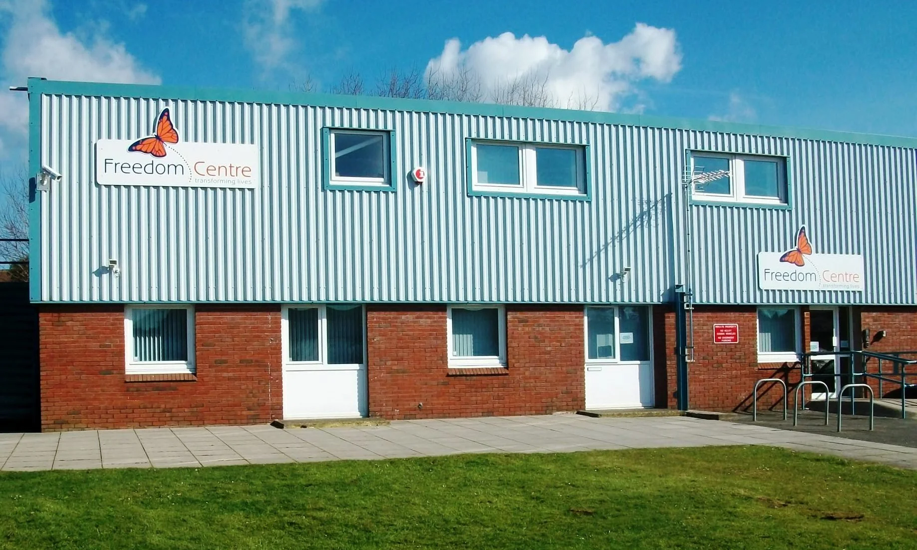 A two-story building with a brick lower level and a corrugated metal upper level. Signs on the building read ‘Freedom Centre’ with a butterfly logo. The building has several windows and a sidewalk in front with a grassy area.