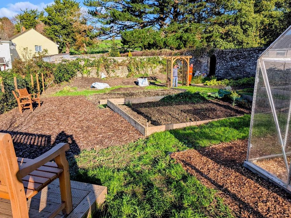 A backyard garden with raised garden beds, a greenhouse, and a person near a wooden archway, surrounded by trees and a stone wall.
