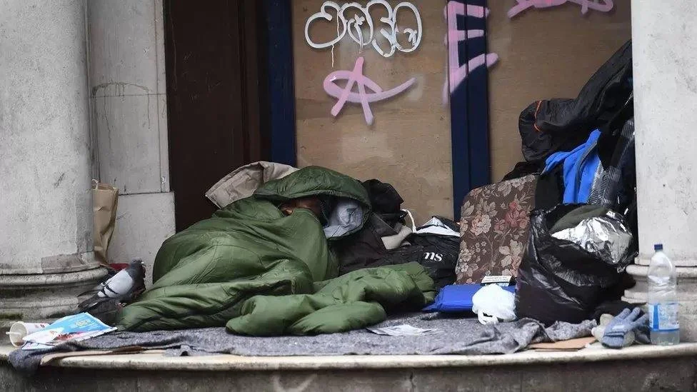A person sleeping on a cardboard on a sidewalk near a wall covered with graffiti, surrounded by trash and bags.
