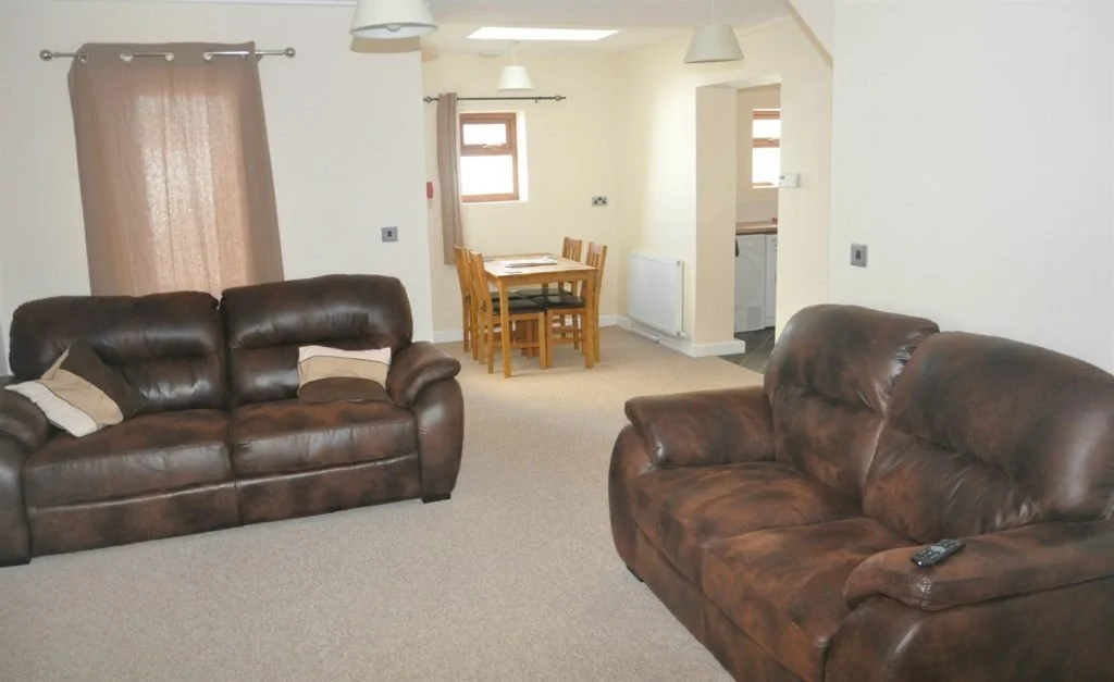 Living room with two brown leather couches, a wooden dining table with four chairs, beige curtains, three ceiling lights, and a doorway leading to the kitchen.