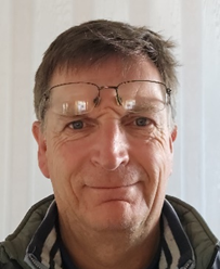 Close-up of a middle-aged man with short brown hair, glasses on his forehead, smiling at the camera, wearing a dark jacket with a striped shirt underneath.