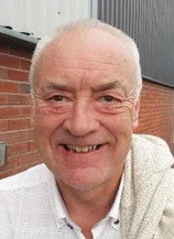A smiling older man with a bald head and light skin, wearing a white collared shirt, standing outdoors in front of a brick wall and gray siding.