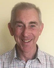 An older man with short gray hair, wearing a checkered shirt, smiling against a plain background.