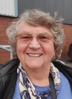A smiling elderly woman with curly gray hair and glasses standing outdoors in front of a brick building and metal siding.
