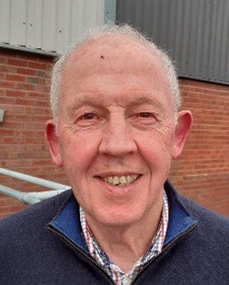 Close-up of an elderly man smiling outdoors, with a brick building and gray wall in the background.