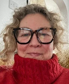 A woman with curly blonde hair wearing large black glasses and a red turtleneck.