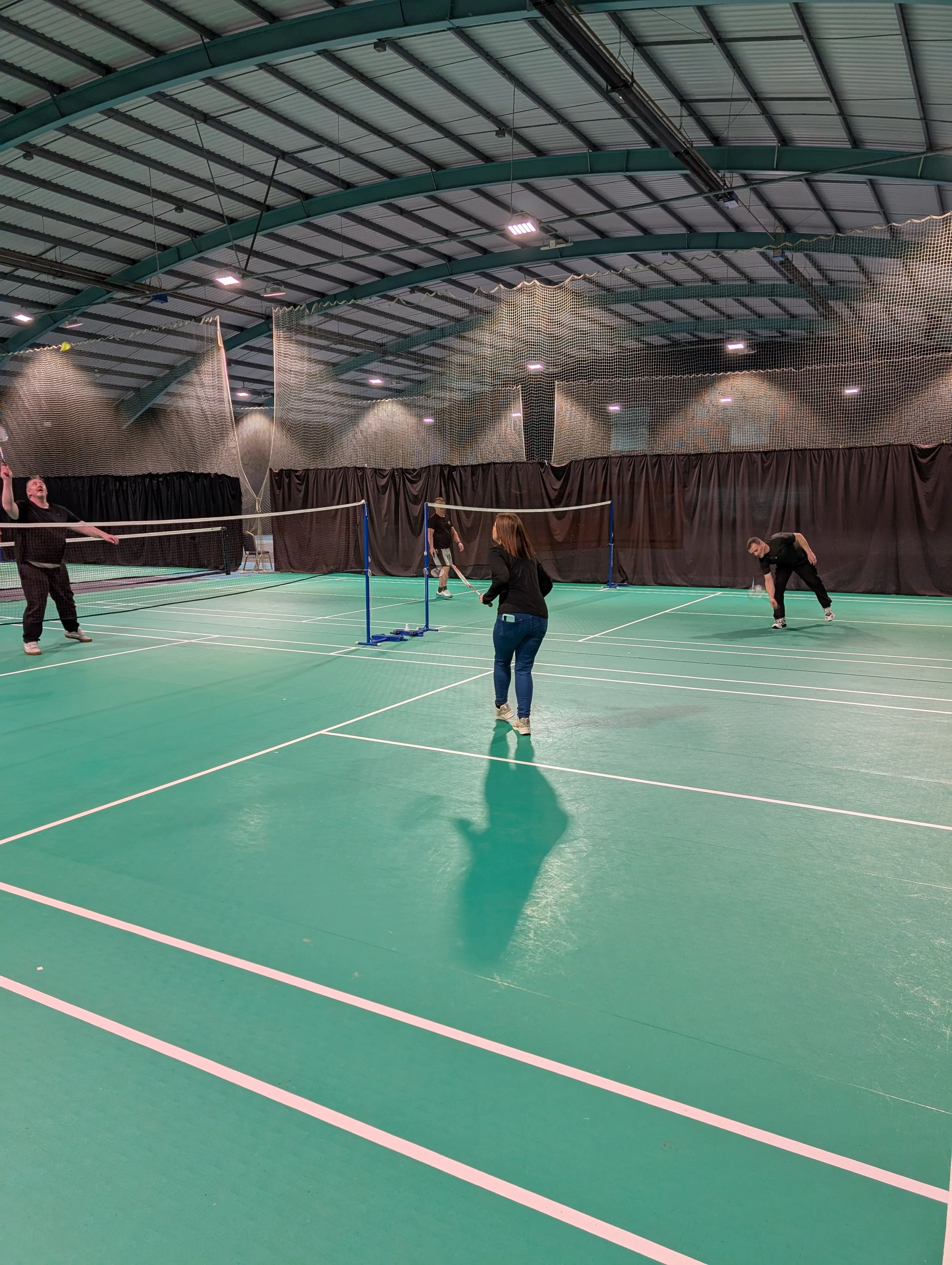 People playing pickleball on an indoor court with green flooring and a high arched ceiling.