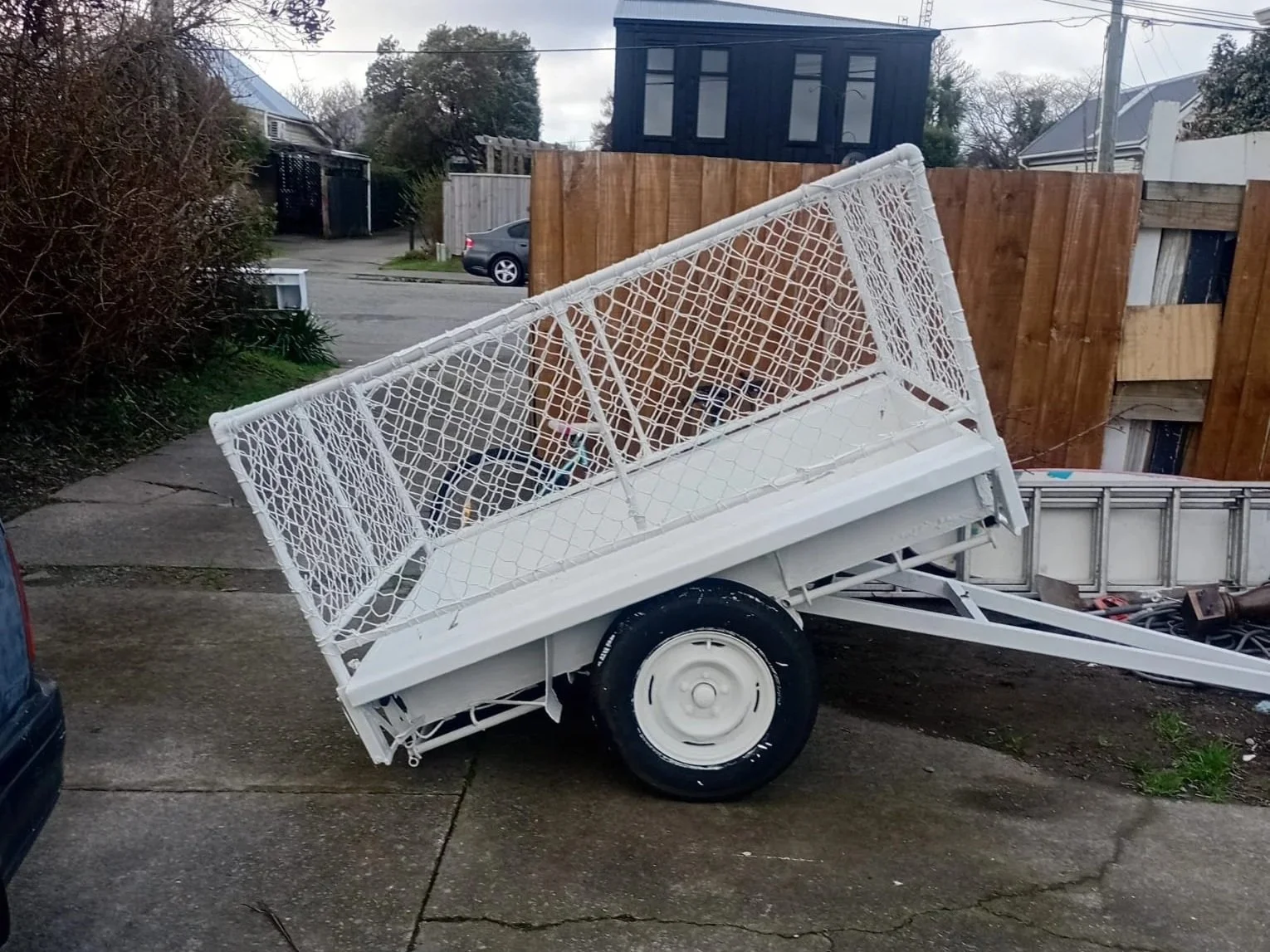 White utility trailer with a tilted mesh sides, parked on a concrete driveway, adjacent to a black vehicle, with trees, houses, and a wooden fence in the background.