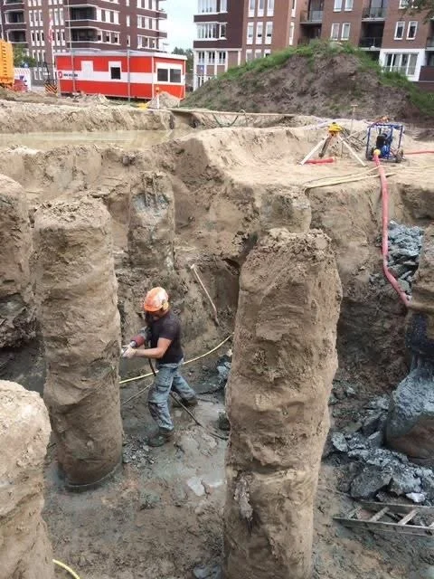 Een bouwplaats met een dieptegraf waar een man in een installatie met een helm en werkoutfit werkt, omringd door grote zand- en rotsformaties.