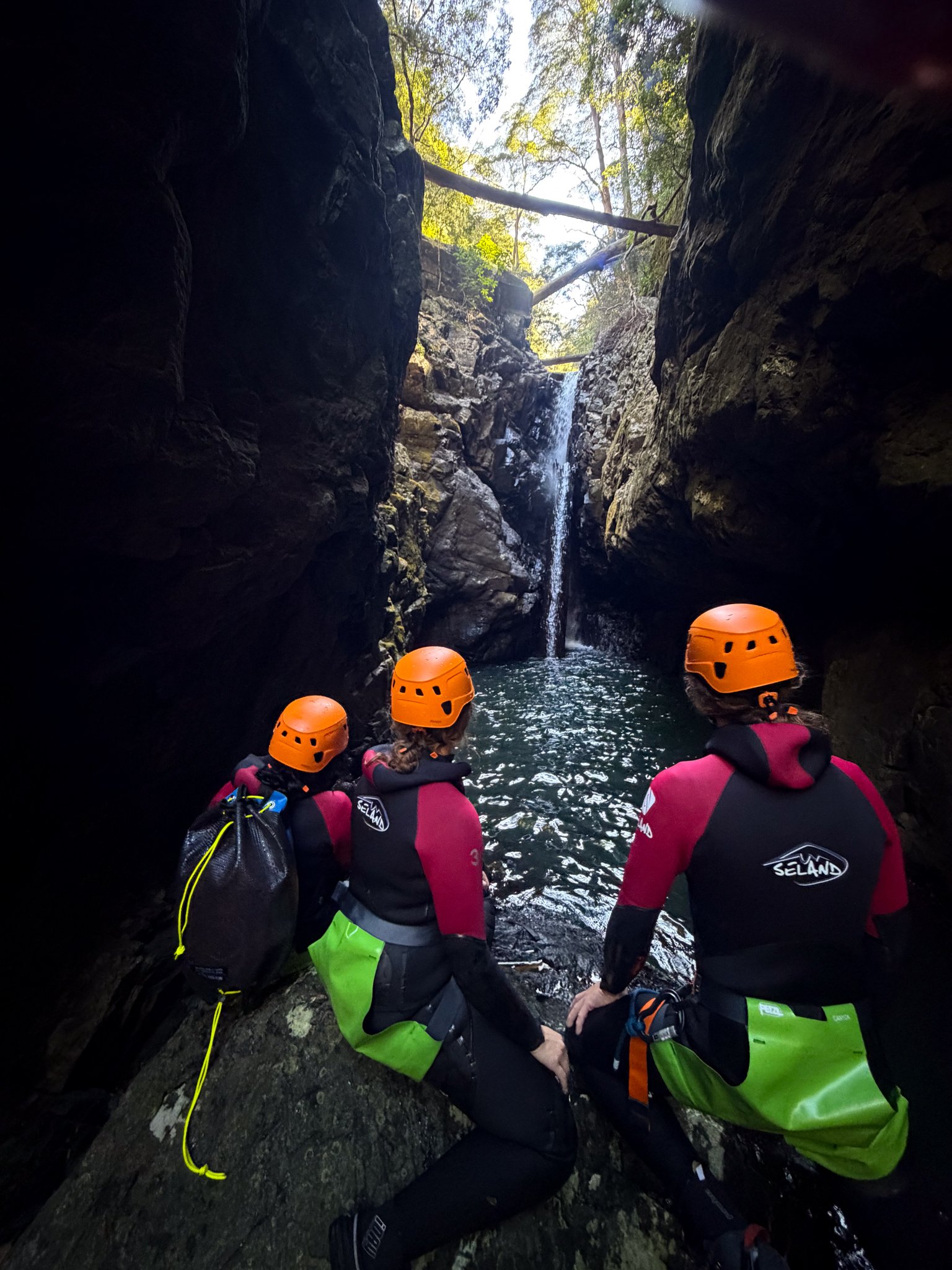 Canyoners in orange helmets looking up through a narrow gorge towards a waterfall.