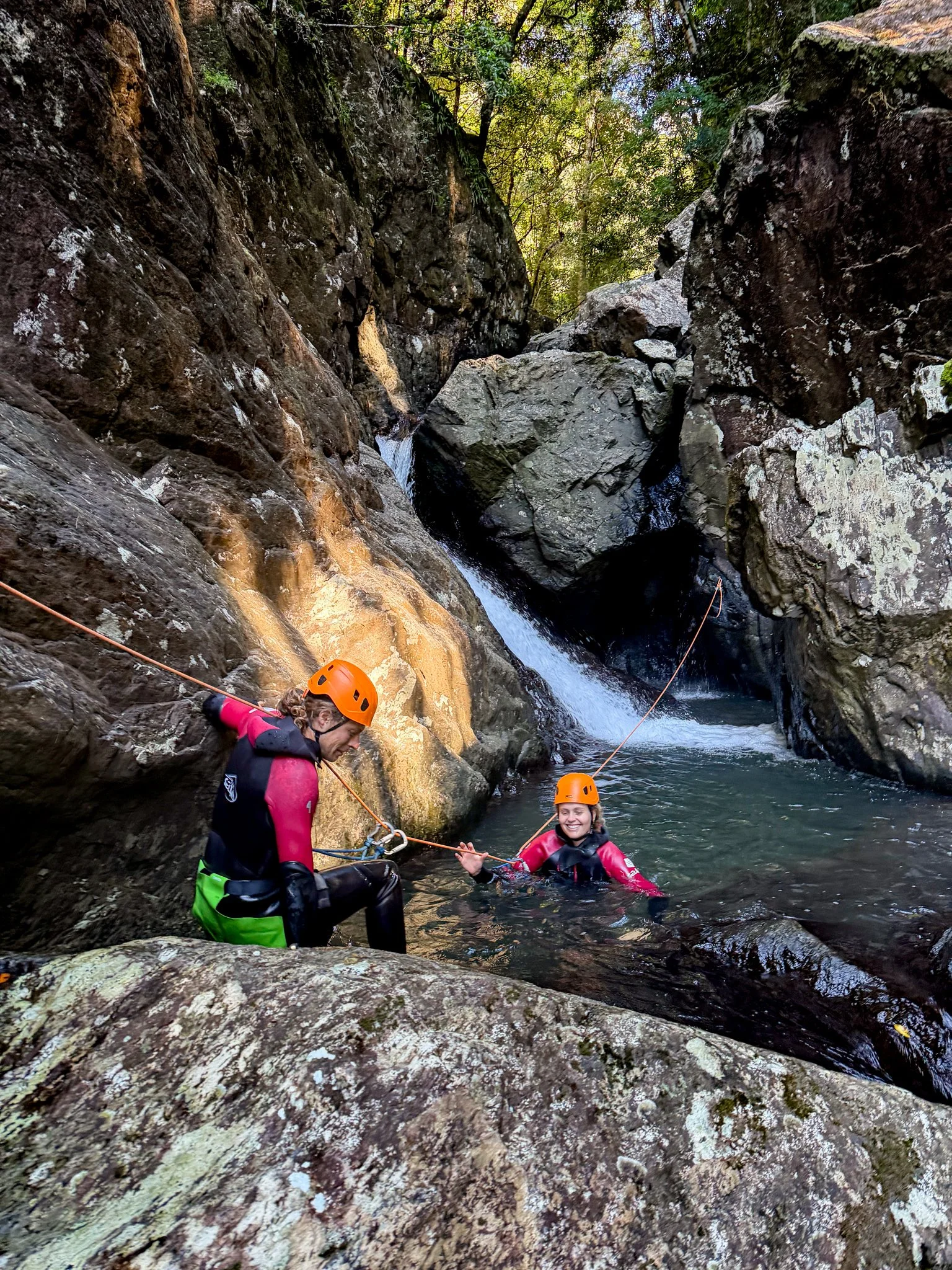 Two canyoners in orange helmets beside a waterfall and rocky pool.