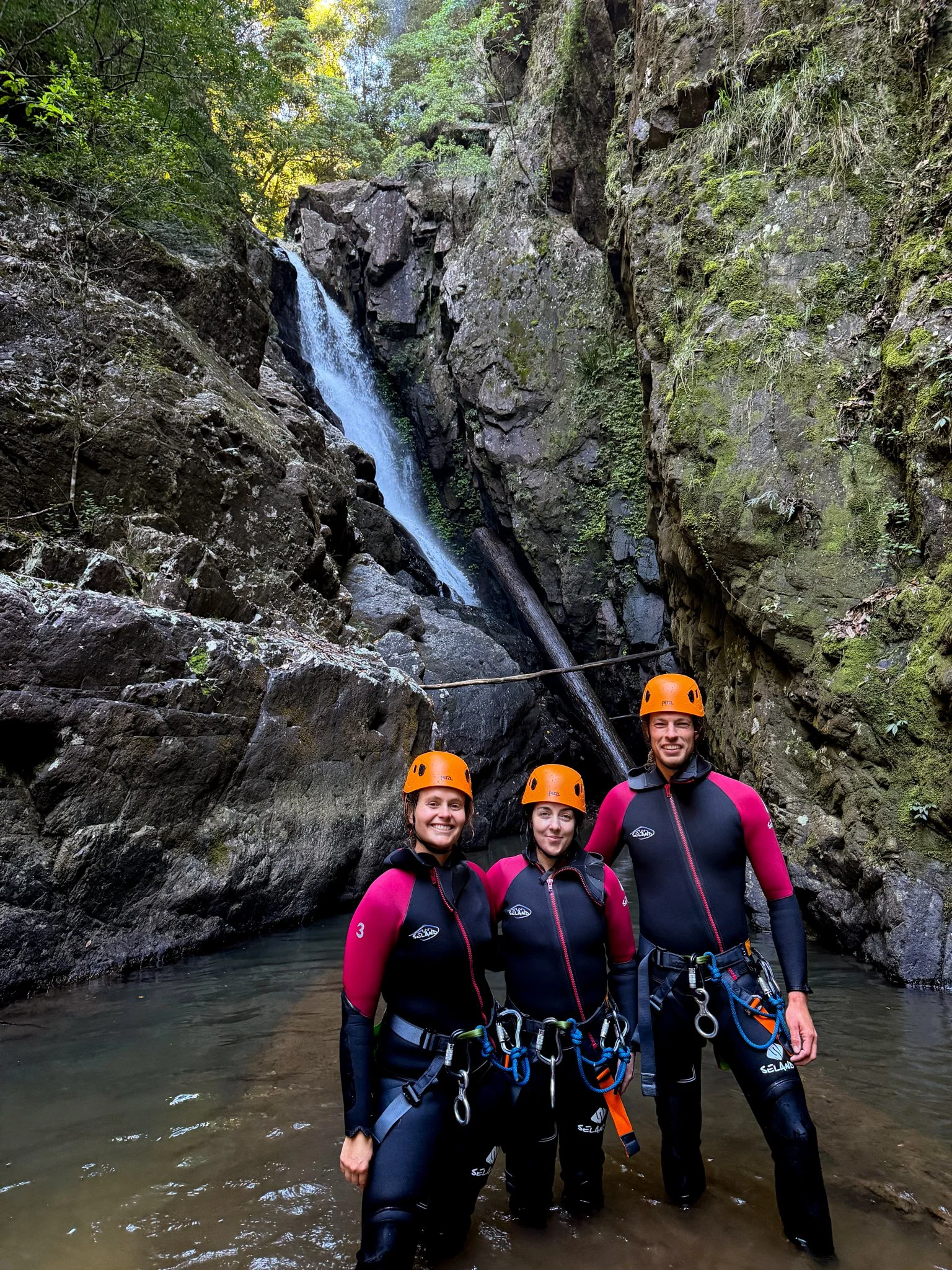 Canyoners in wetsuits and helmets standing below a waterfall in a rocky gorge.