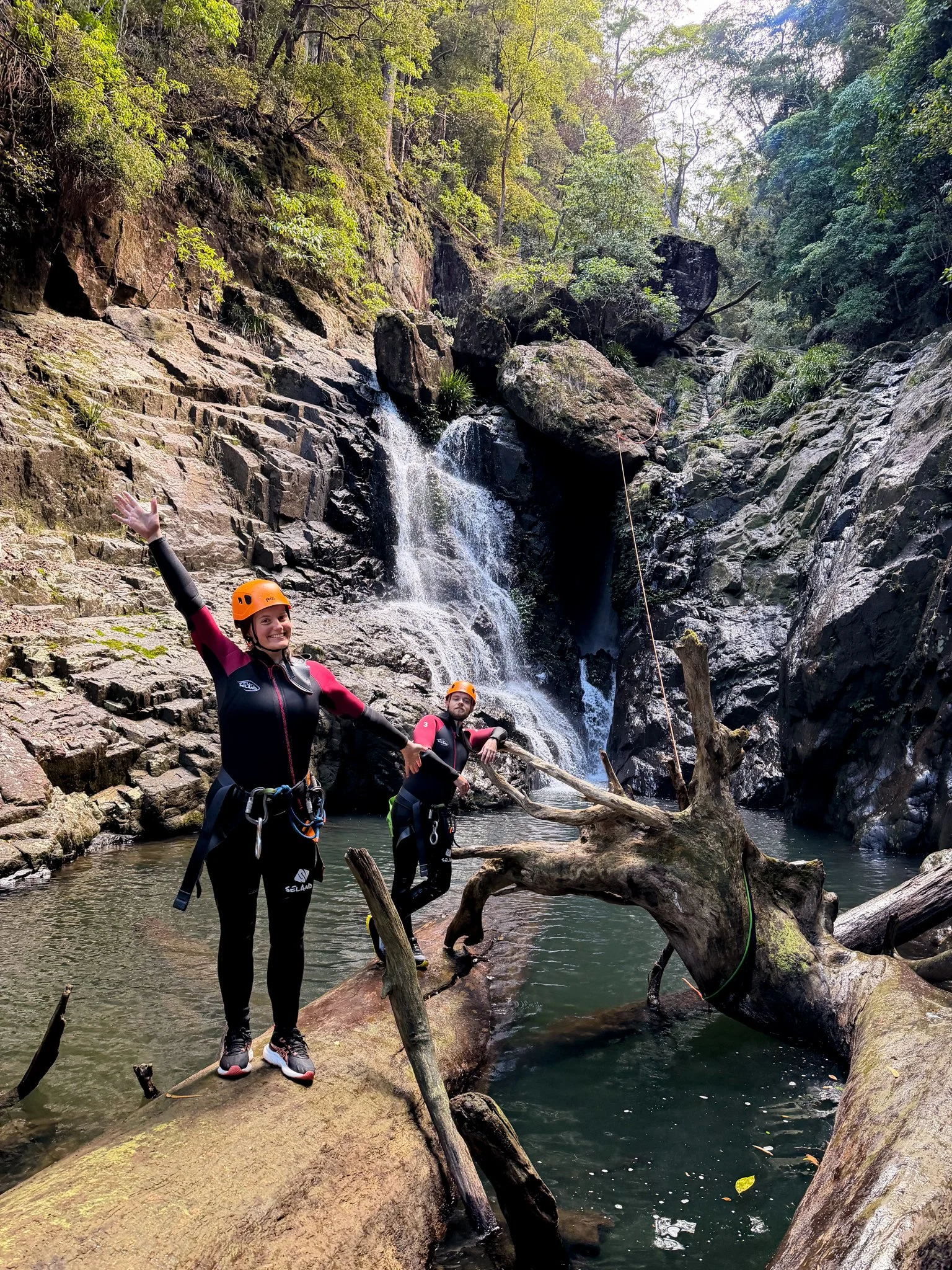 Two people in wetsuits and helmets standing on rocks in a river with a waterfall and lush green trees in the background.