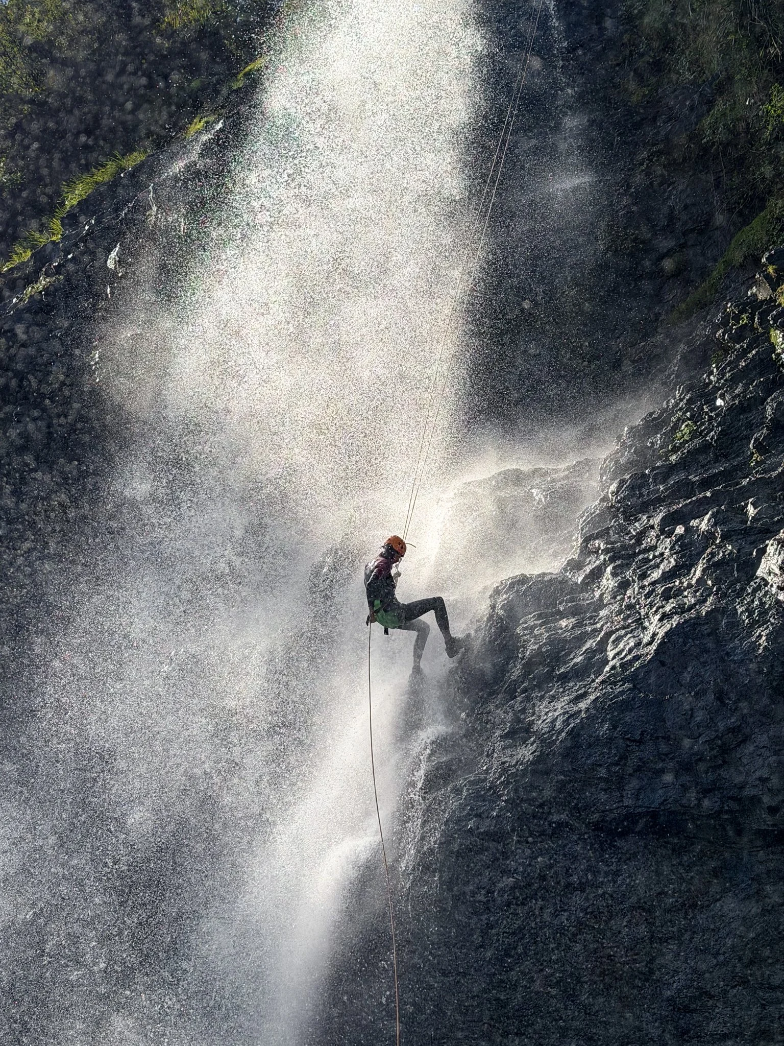 Canyoner abseiling through the spray of a waterfall on a steep rock face.