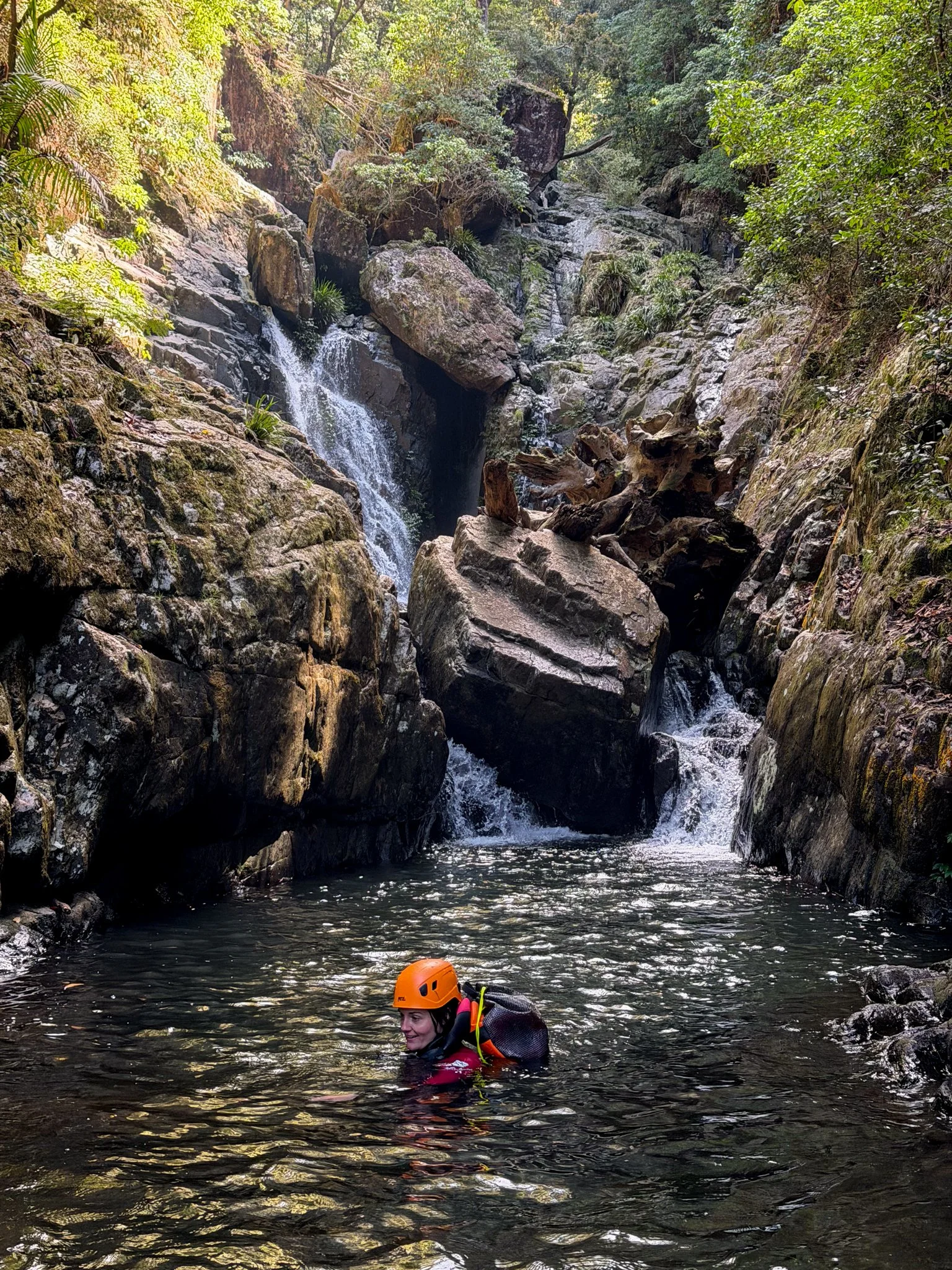 A person wearing an orange helmet and black life jacket swimming in a river inside a rocky, forested canyon with a small waterfall.