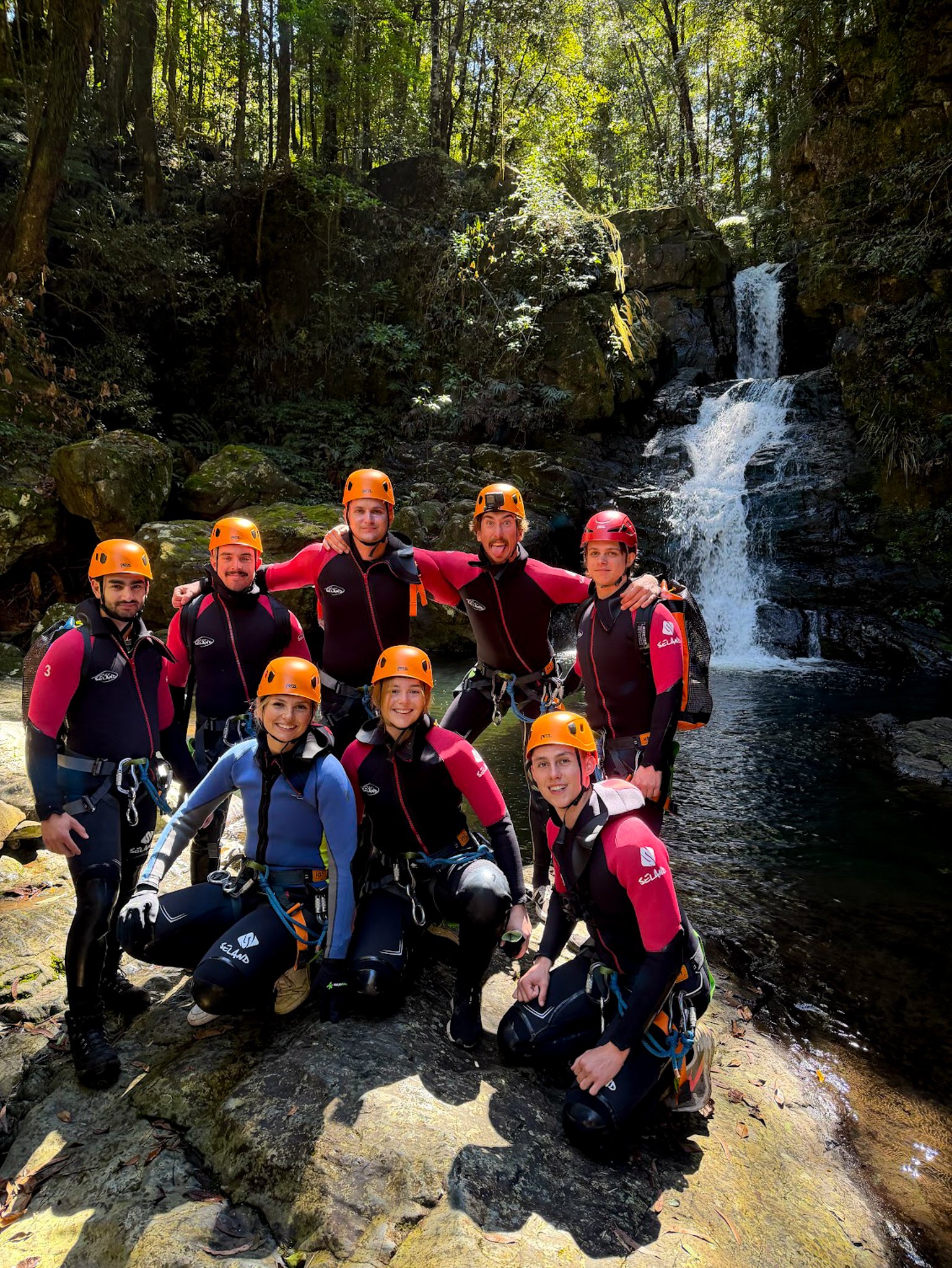 Group of people in outdoor gear with helmets, standing by a small waterfall in a forested area.