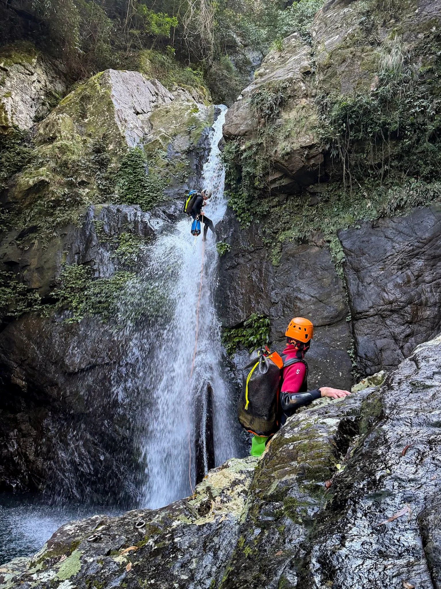 Canyoner in an orange helmet beside a waterfall between steep rock walls.
