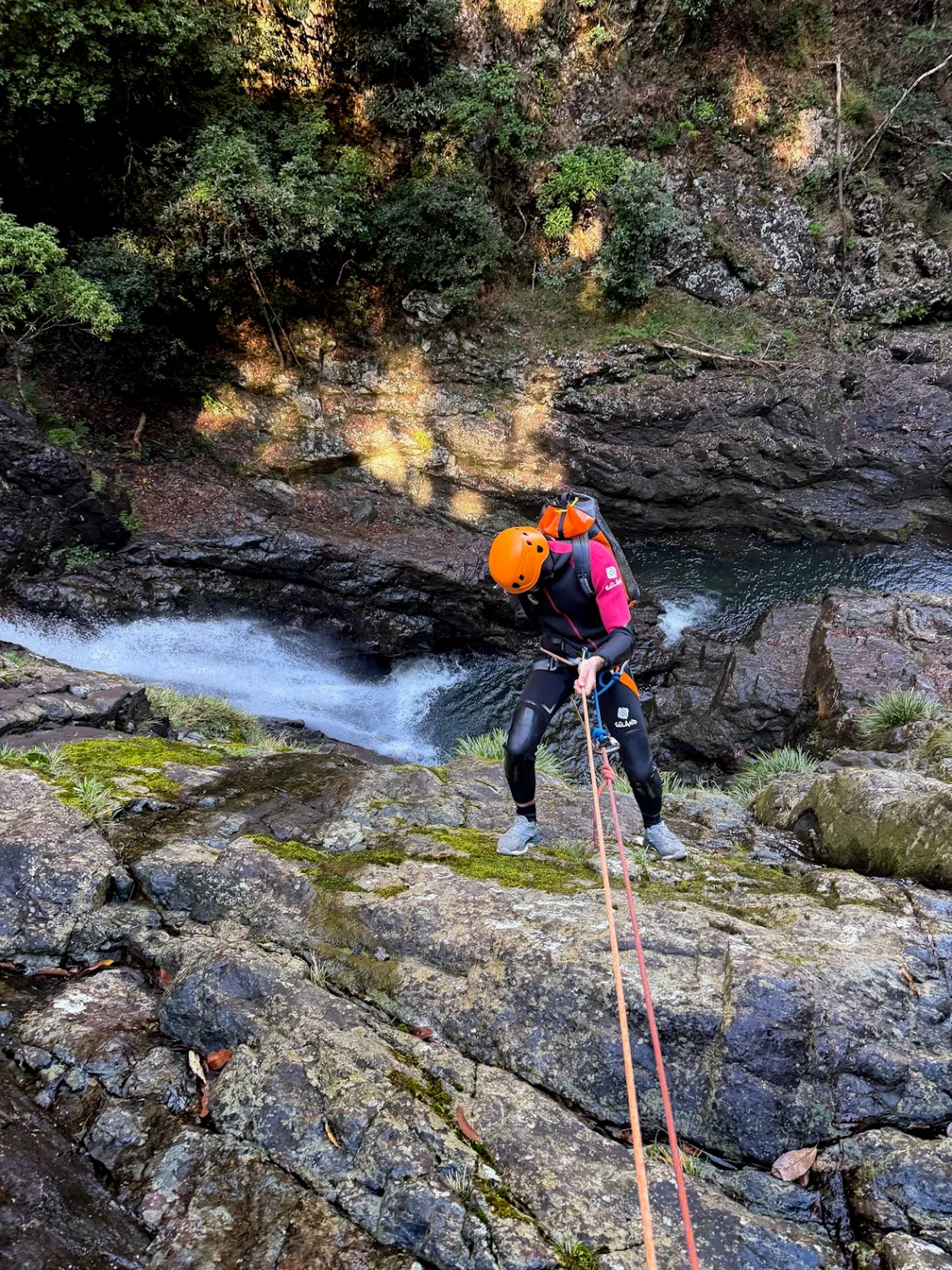Canyoner in an orange helmet using a rope on rocks beside a rainforest stream.