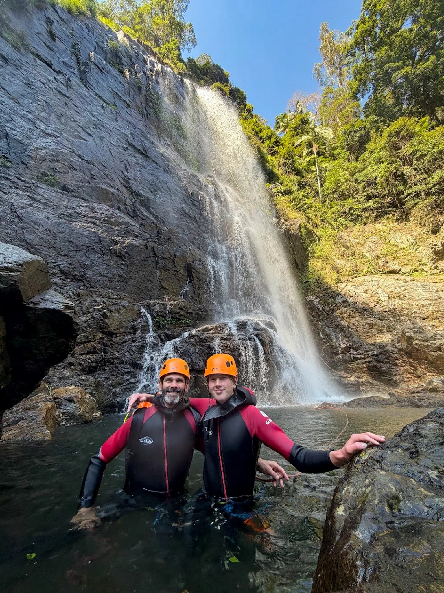Two canyoners in orange helmets at the base of a tall waterfall.