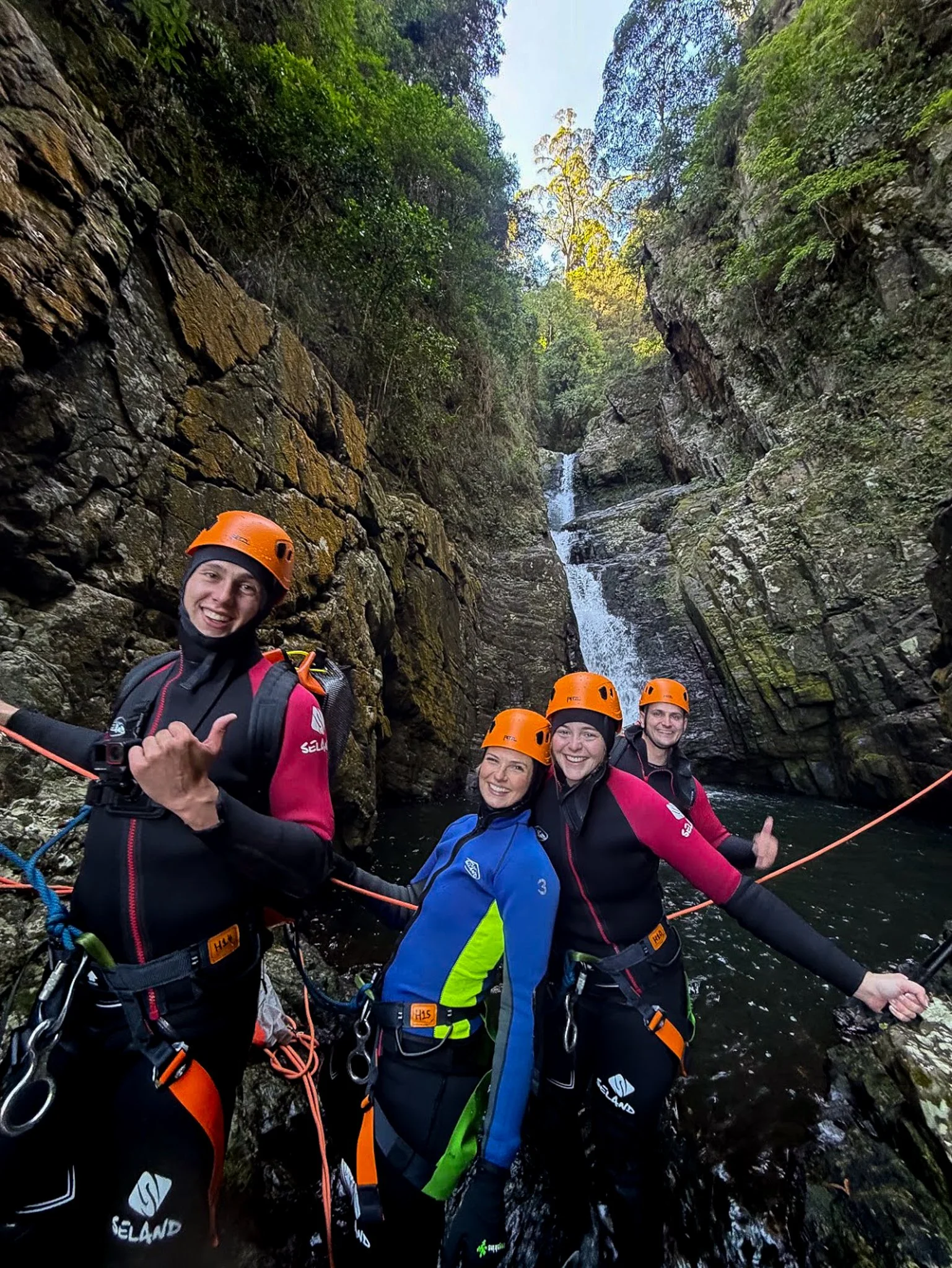 Group of people in wetsuits and helmets standing in a rocky canyon with a waterfall in the background, smiling and posing for the camera during a canyoning adventure.