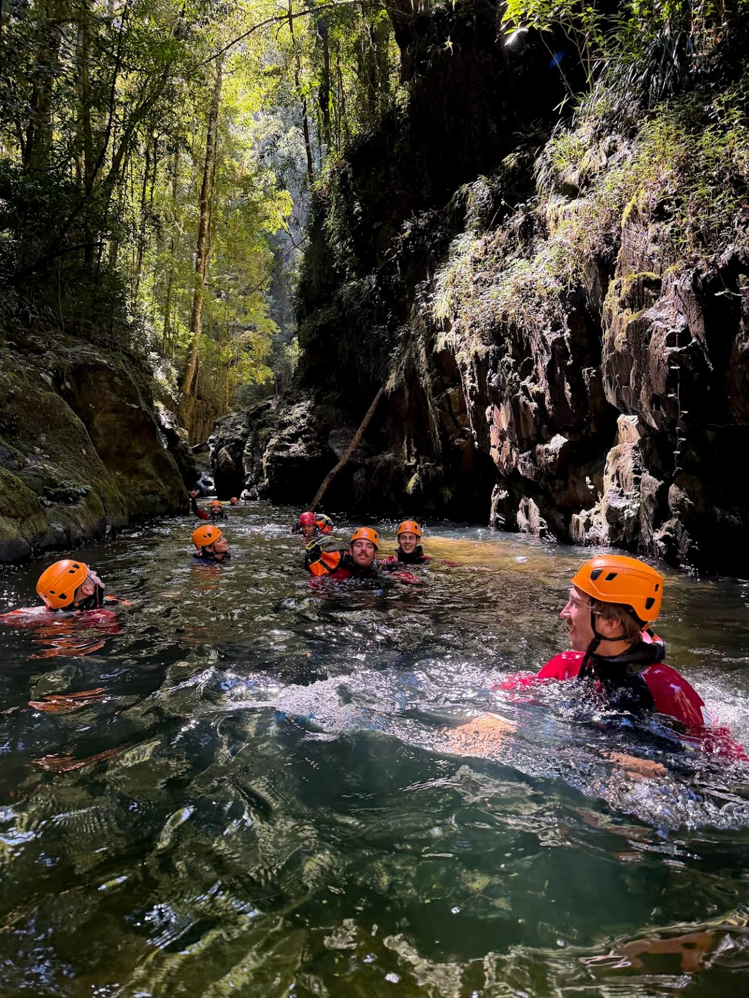 Canyoners in orange helmets swimming through a narrow rocky gorge.