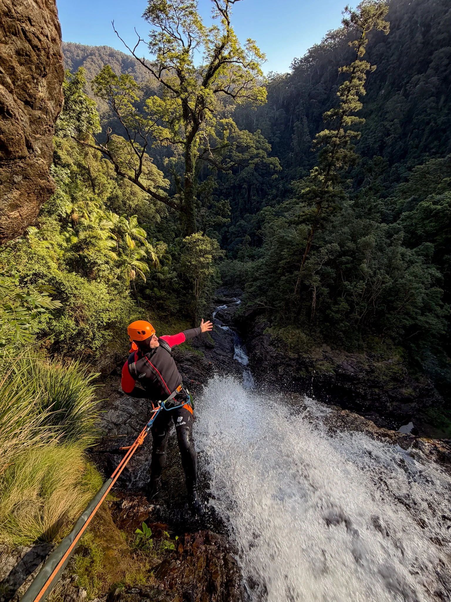 Canyoner wearing a helmet and harness beside flowing water in a rainforest gorge.