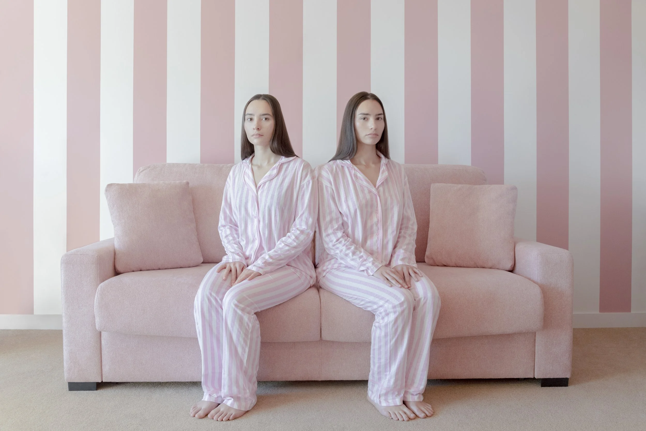 Two women with long dark hair sitting on a pink couch with pink and white striped pajamas, in a room with pink and white vertically striped wallpaper and beige carpet.